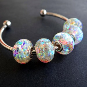 May include: A silver-toned cuff bracelet featuring six iridescent, faceted beads. The beads have a clear base with colorful, glitter-like inclusions. The bracelet has a simple, curved design with small, round end caps.