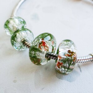 Peut inclure: Un bracelet à breloques argenté avec quatre perles en verre transparent. Chaque perle contient des feuilles vertes séchées et de petites fleurs rouges et orange.