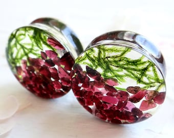 May include: A pair of clear, round ear plugs with a botanical design. The plugs are filled with small, red stones and green plant matter, encased in clear resin. The plugs have a shiny, metallic border.