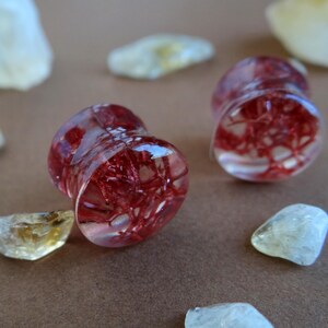 May include: Two clear red and white resin ear gauges with a red, web-like pattern inside.