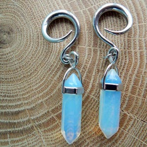 May include: A pair of silver ear gauges with a dangling opal crystal charm. The gauges are made of metal and have a spiral design. The opal crystals are a light blue color.