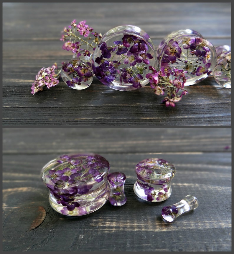 May include: A set of clear resin ear gauges with purple and pink pressed flowers. The gauges are different sizes and are displayed on a dark wooden surface.