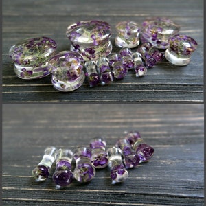 May include: A set of clear acrylic ear gauges with purple pressed flowers inside. The gauges are in various sizes and are arranged on a dark wooden surface.