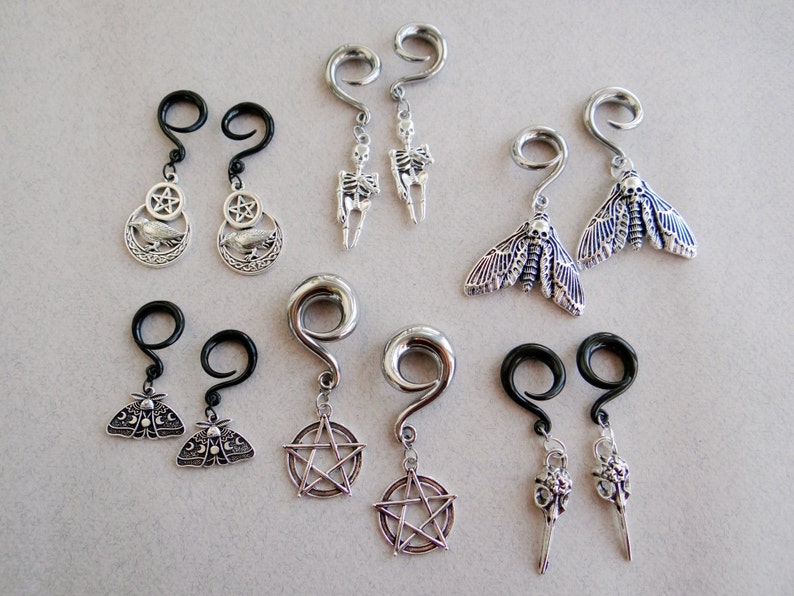 May include: A set of eight silver and black dangle earrings with various charms, including a skull, a moth, a pentagram, and a crow. The earrings are designed for stretched earlobes.