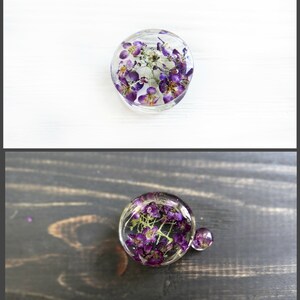 May include: Two round resin ornaments with purple flowers pressed inside. The ornaments are clear and show the flowers in detail. The larger ornament is on a dark wood surface, while the smaller ornament is on a white surface.