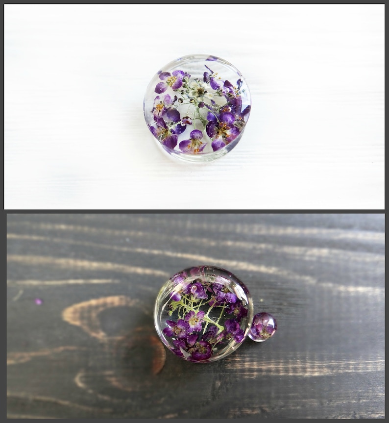 May include: Two round resin ornaments with purple flowers pressed inside. The ornaments are clear and show the flowers in detail. The larger ornament is on a dark wood surface, while the smaller ornament is on a white surface.