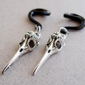 May include: A pair of silver bird skull earrings with black rubber ear cuffs. The earrings are designed to look like the skull of a bird with a long beak.