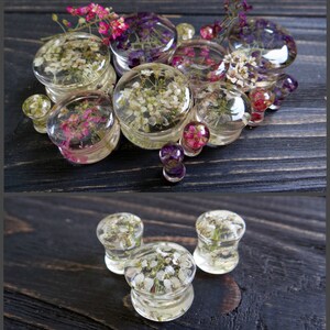 May include: Assortment of clear resin ear gauges with embedded dried flowers. The gauges are various sizes and feature different flower arrangements, including white, pink, and purple flowers.
