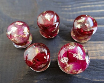 May include: Five round, clear, resin ear gauges with embedded dried rose petals and gold leaf. The gauges are various shades of pink and red.