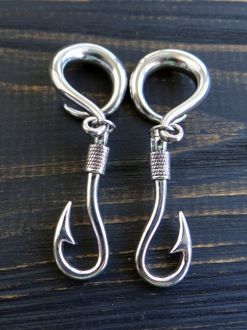 Op de afbeelding: Een paar zilveren metalen oorpluggen met een vishaakontwerp. De pluggen zijn rond en hebben een gladde afwerking.