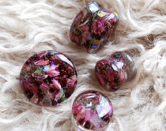 May include: Four resin objects containing dried purple flowers. The shapes are a circle, a heart, a cube, and a small sphere. The objects have a clear, glossy finish. They are displayed on a fluffy, white surface.