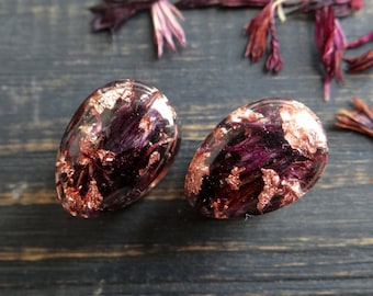 May include: A pair of oval-shaped earrings with a clear resin base, filled with dried purple flowers and copper flakes.
