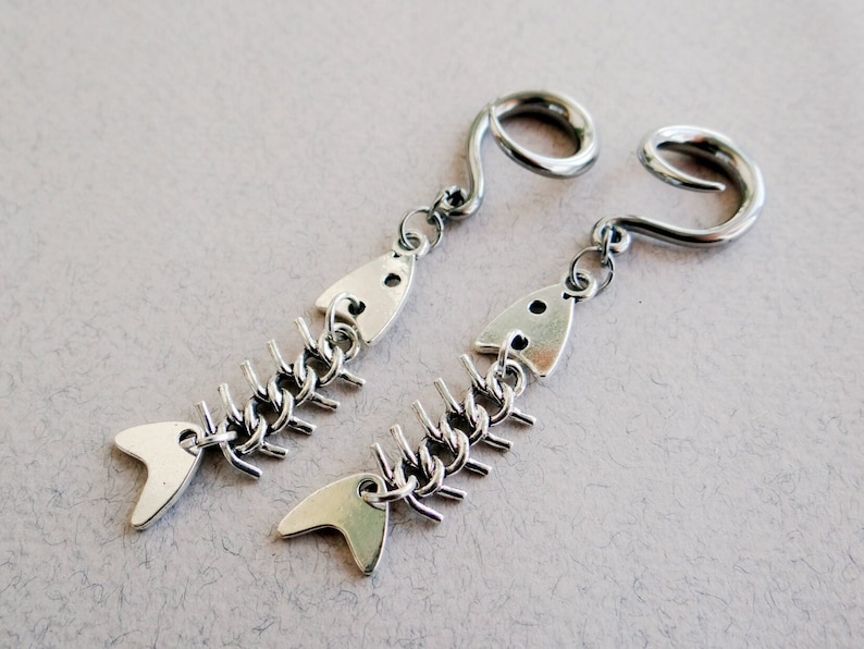 May include: A pair of silver fishbone earrings with a dangling charm. The earrings are made of metal and have a silver finish. The fishbone charm is detailed and realistic.