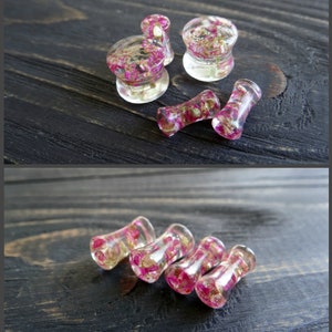 May include: A set of six clear acrylic ear gauges with pink and gold flower accents. The gauges are in various sizes and shapes, including round, cylindrical, and flared.