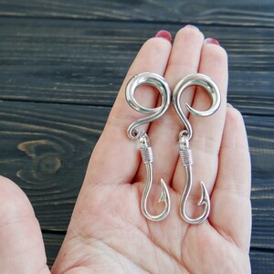 Op de afbeelding: Een paar zilveren vishaak oorbellen. De oorbellen hebben de vorm van een vishaak met een ronde bovenkant. De oorbellen hangen aan de hand van een persoon.