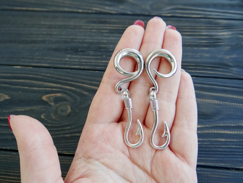 Op de afbeelding: Een paar zilveren vishaak oorbellen. De oorbellen hebben de vorm van een vishaak met een ronde bovenkant. De oorbellen hangen aan de hand van een persoon.