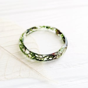 Puede incluir: Un anillo de resina transparente con follaje verde y marrón incrustado. El anillo está sobre una superficie blanca.