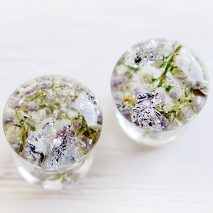 Può includere: Due tappi rotondi in resina trasparente con fiori secchi e foglia d'argento incorporati. I fiori sono bianchi e viola.