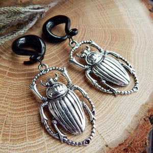 May include: A pair of black horn ear gauges with silver beetle charms. The beetles have detailed wings and legs.