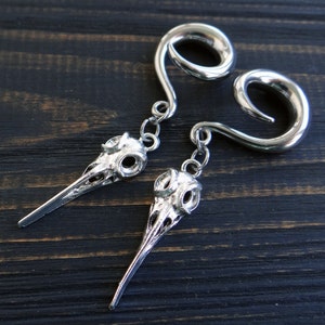 May include: A pair of silver metal ear gauges with a skull design hanging from each. The gauges are shaped like a spiral and the skulls have a bird-like beak.