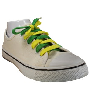 Puede incluir: Una zapatilla de lona de corte bajo en color blanco roto con puntera de goma blanca y una raya negra a lo largo de la suela. El calzado presenta cordones planos que alternan el verde brillante y el amarillo, creando un patrón multicolor.
