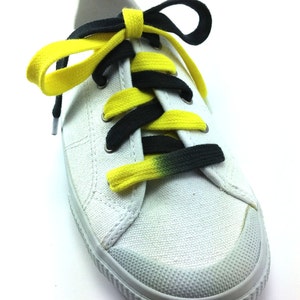 Puede incluir: Una zapatilla de lona blanca con puntera de goma texturizada y cierre de cordones. El calzado presenta cordones planos que transicionan de negro sólido a amarillo brillante, creando un patrón de alto contraste sobre la tela blanca superior.