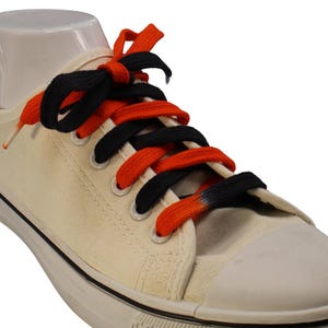 Puede incluir: Zapatillas blancas de lona con cordones negros y naranjas. Los cordones están atados en forma de cruz.