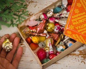 May include: A box filled with vintage glass Christmas ornaments in various colors and shapes, including red, gold, silver, and clear. The box is labeled in Russian with the words "Elochnye ukrasheniya Maliotka" which translates to "Christmas tree decorations Little One".