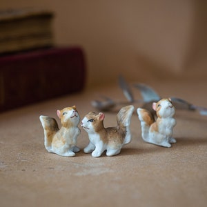 May include: Three small ceramic squirrel figurines. The squirrels are white, with brown and tan markings. They have upright tails and are posed in different positions. The background includes a blurred book and dried foliage.
