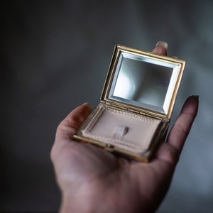 Op de afbeelding: Een vintage goudkleurige compacte spiegel, open om een spiegel en een poedercompartiment met een beige poederdons te onthullen. De compact heeft een rechthoekige vorm met een scharnierend deksel.
