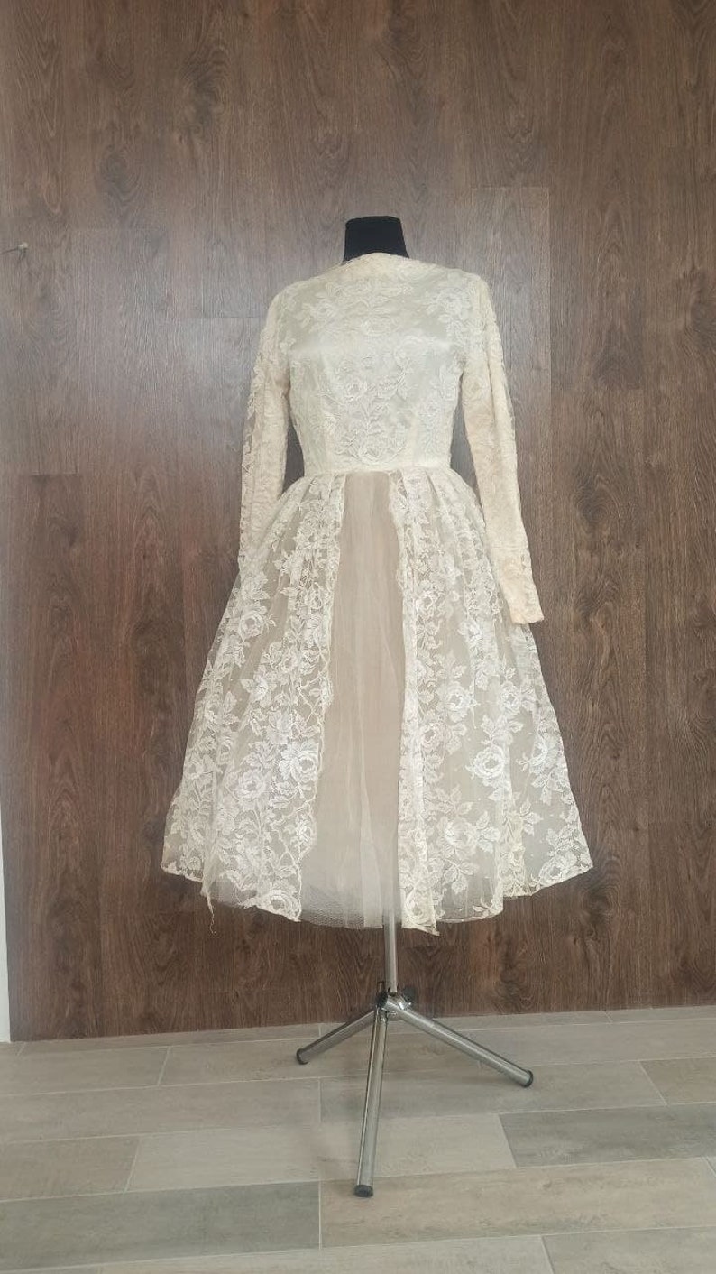 Op de afbeelding: Ivoor vintage jurk met lange mouwen en een hoge halslijn. De jurk heeft een bloemen kanten overlay over een lichter gekleurde onderjurk. De jurk wordt getoond op een mannequin tegen een achtergrond van houten panelen.