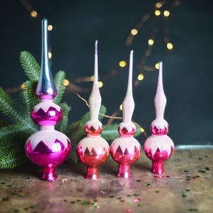 May include: Four vintage-style glass Christmas tree toppers in varying sizes. Each ornament features a shiny magenta base with a pointed spire, accented by a matte, frosted white finish on the upper sections and a distinct zigzag pattern.