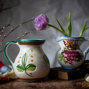 May include: Two ceramic jugs with floral designs. One jug has a green handle and leaf design, the other has a grape design and holds purple tulips. The jugs sit on a wooden surface.