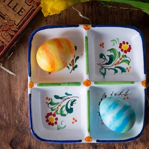 Puede incluir: Un plato cuadrado dividido de cerámica con borde azul, con dos huevos de Pascua decorados. El plato está decorado con motivos florales en verde, rojo y naranja. Un libro y flores están en el fondo.