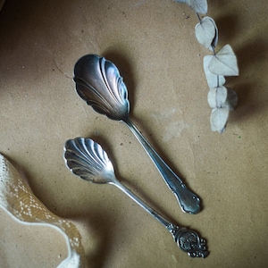 May include: Two silver spoons with ornate handles and a scalloped design. The spoons are on a brown surface.