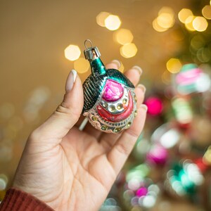 May include: A vintage glass Christmas ornament with a green, silver, and pink design. The ornament is shaped like a bell and has a silver loop for hanging.