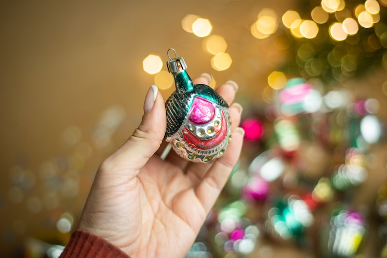 May include: A vintage glass Christmas ornament with a green, silver, and pink design. The ornament is shaped like a bell and has a silver loop for hanging.