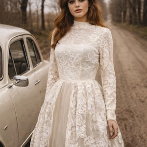 Op de afbeelding: Cr&egrave;mekleurige kanten trouwjurk met lange mouwen en een hoge halslijn. De jurk heeft een volle rok en gedetailleerd bloemenkant. Een witte haarband wordt gedragen. Een oldtimer staat op de achtergrond.