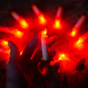 Peut inclure: Une guirlande de bougies électriques émettant une lueur rouge vive, disposées en cercle. Chaque bougie présente un corps cylindrique blanc à la texture cireuse, surmonté d'une petite ampoule en forme de flamme illuminée sur fond sombre.