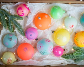 May include: A collection of round glass Christmas ornaments in various colors including orange, yellow, pink, light blue, and green. Each ornament features painted floral or geometric patterns and a silver metal cap. Green pine branches are visible.