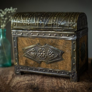 May include: A vintage wooden chest with metal accents. The chest has a domed lid and is adorned with decorative metalwork. The wood has a natural grain, and the metal has a tarnished appearance. The chest is on a wooden surface.