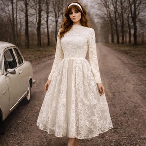 Op de afbeelding: Een cr&egrave;mekleurige jurk met lange mouwen, een hoge halslijn en een wijde rok. De jurk is gemaakt van kant en heeft een vintage stijl. Een witte haarband wordt gedragen. Een oldtimer staat in de buurt.