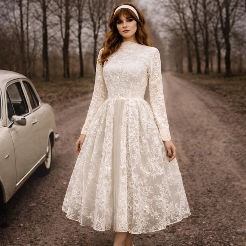 Op de afbeelding: Een cr&egrave;mekleurige jurk met lange mouwen, een hoge halslijn en een wijde rok. De jurk is gemaakt van kant en heeft een vintage stijl. Een witte haarband wordt gedragen. Een oldtimer staat in de buurt.