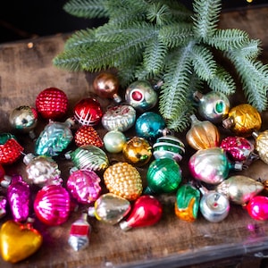 May include: A collection of vintage Christmas ornaments in various shapes and colors, including red, pink, gold, and green. Some ornaments have textured surfaces, and a sprig of evergreen is in the background. Holiday decor.