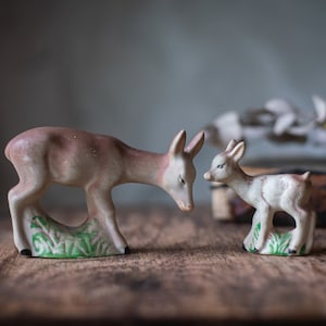 May include: Two ceramic deer figurines. One is larger, with a light brown body and a white face, looking down at a smaller fawn. Both stand on green grass bases. The figurines are on a wooden surface.