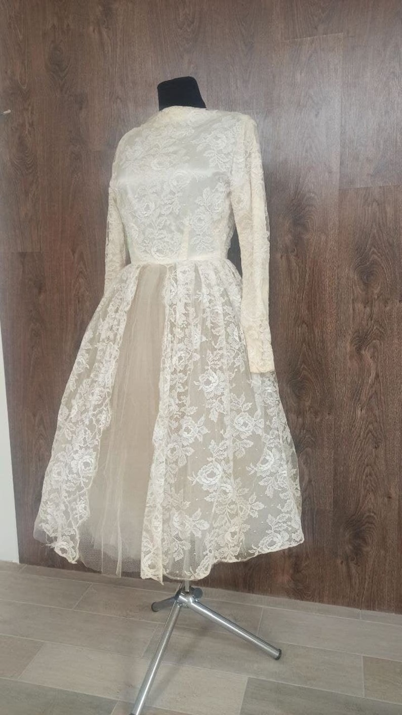 Op de afbeelding: Een vintage ivoorkleurige kanten jurk met lange mouwen en een hoge halslijn. De jurk heeft een bloemen kanten overlay en een tule rok, weergegeven op een paspop. De jurk is knielang en heeft een klassieke, elegante stijl.