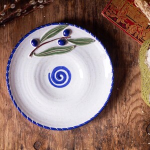 May include: White ceramic plate with a blue spiral design in the center and a blue rim. The plate is decorated with a painted olive branch with green leaves and blue olives. The plate is on a wooden surface.