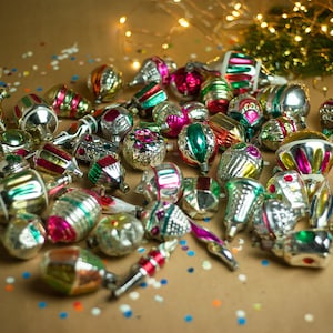 May include: A collection of vintage glass Christmas ornaments in various colors and designs, including silver, gold, red, green, and pink. The ornaments are scattered on a brown surface with confetti.