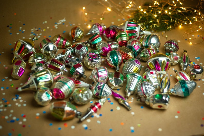 May include: A collection of vintage glass Christmas ornaments in various colors and designs, including silver, gold, red, green, and pink. The ornaments are scattered on a brown surface with confetti.
