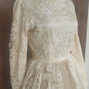 Op de afbeelding: Ivoor kanten trouwjurk met lange mouwen en een hoge halslijn. De jurk heeft ingewikkelde bloemenkanten details over de hele bodice en rok, waardoor een romantische en elegante look ontstaat. De rok heeft een vloeiend, gelaagd ontwerp.